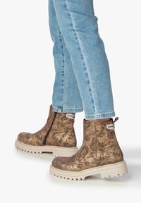 Bottines métalliques brunes avec une finition texturée, fermeture éclair sur le côté et semelle en caoutchouc clair. Portées avec un jean bleu, montrant les jambes inférieures.