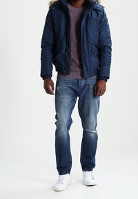 Doudoune marine avec une capuche bordée de fourrure, pull en tricot bordeaux, jean bleu déchiré et baskets blanches. Tenue décontractée superposée.