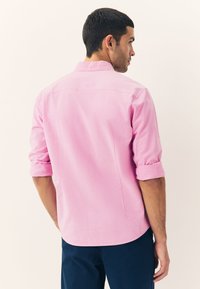Light pink button-up shirt with rolled sleeves, made of a smooth fabric. Features a back yoke and side slits for ease of movement.