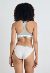 Šedý športový podprsenka a biele nohavičky od značky Calvin Klein, s logom na elastickom páse, hladkým povrchom a dizajnom s prekríženými ramienkami. Žiadne vzory nie sú viditeľné.