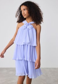 Robe lilas claire plissée avec des volants superposés, ornement floral au niveau du décolleté, et un design à col licou. Texture de tissu douce et fluide.