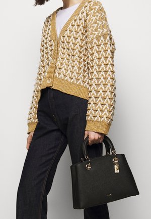 Cardigan giallo e bianco con motivo, scollatura a V, abbinato a jeans scuri. Tiene in mano una borsa nera con dettagli dorati e un portachiavi.