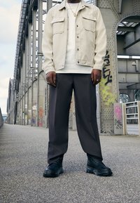 Chaqueta beige claro con dos bolsillos frontales sobre una camisa blanca, combinada con pantalones anchos gris oscuro y botas negras gruesas. Fondo urbano.