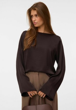 Mujer con pelo largo y castaño que lleva una blusa suelta de manga larga de color marrón oscuro y pantalones beige, sosteniendo un bolso marrón delante de ella.