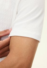 Camicetta bianca di cotone a maniche corte con un tessuto testurizzato sul corpo e polsini delle maniche lisci. Ingrandimento che mostra l'adattamento del tessuto sull'avambraccio.