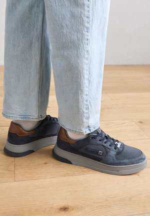 Person trägt hellblaue Jeans und dunkelgraue Turnschuhe mit braunen Akzenten und steht auf einem Holzboden.