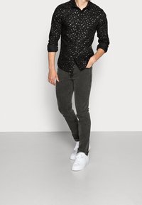 Camicia nera con bottoni e pois dorati e argentati, abbinata a jeans grigio scuro e sneakers bianche, in piedi davanti a uno sfondo neutro.