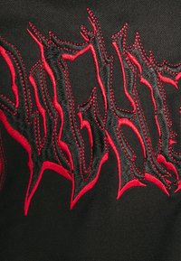 Tessuto nero con audace ricamo rosso frastagliato e un contorno nero a contrasto. Il design ha un aspetto testurizzato e in rilievo.