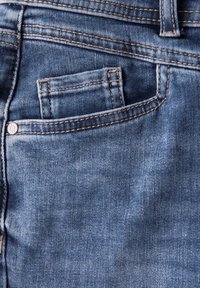 Des jeans en denim bleu présentent une poche arrière avec des détails de couture, un petit bouton en métal, et une texture lisse, légèrement décolorée.