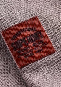 Tessuto grigio con una trama testurizzata presenta un marchio cucito a forma di quadrato di colore rosso con testo in nero in rilievo, "SUPERDRY," e ulteriori dettagli sottostanti.