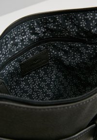 Intérieur d'un sac en cuir noir avec une poche zippée. La doublure en tissu présente un motif floral subtil en gris et blanc sur un fond sombre.