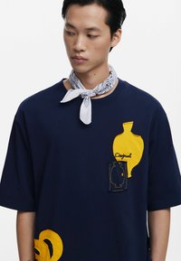 Tamsiai mėlynas per didelis marškinėliai su geltonomis audinio detalėmis, siuvinėtais raštais ir kišenės akcentu. Dėvimi su balta modeliuota bandana.