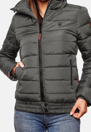 Vrouw draagt een donkergrijze doorgestikte jas met ritssluiting en drukknopen, handen bij de zakken, gecombineerd met blauwe spijkerbroek.