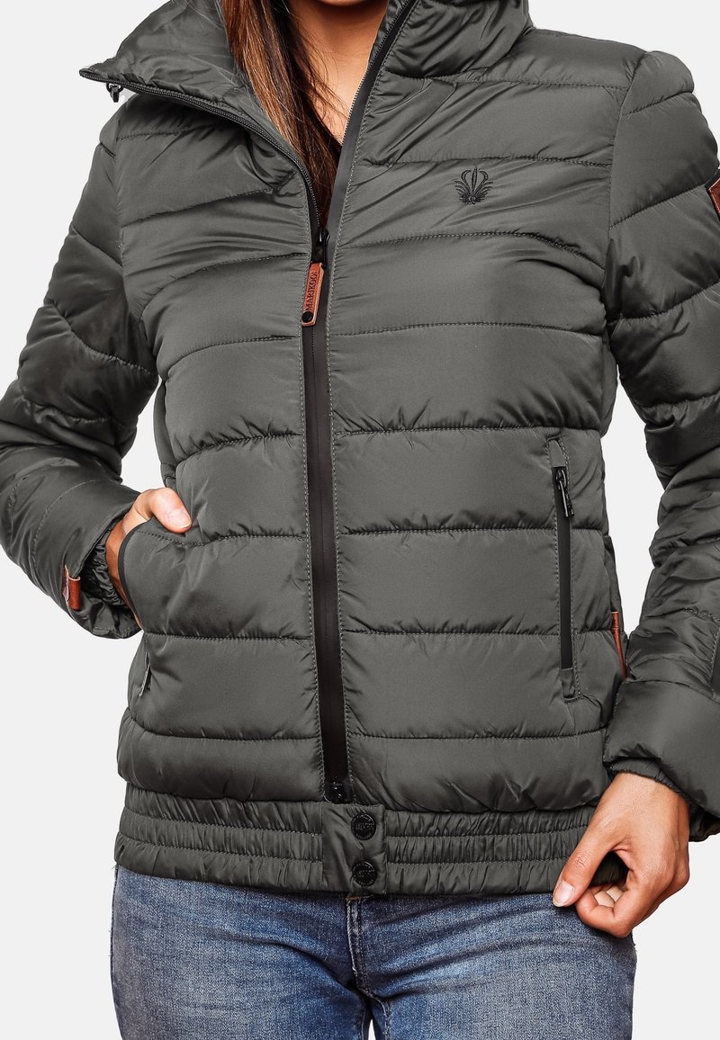 Femme portant une veste matelassée gris foncé avec fermeture éclair et boutons-pression, mains près des poches, associée à un jean en denim bleu.