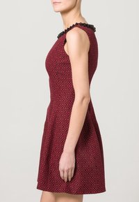 Robe rouge ajustée et évasée avec un col en perles noires, présentant un motif texturé. La robe a un design sans manches et une jupe évasée.