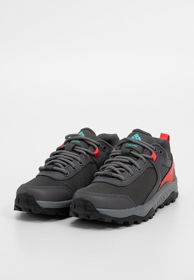 Zapatos de senderismo en negro y gris con tejido de malla, suelas de goma, acentos rojos contrastantes y detalles en azul en el logo. Presentan cordones tradicionales.
