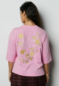 Lyserød bomulds-T-shirt med korte ærmer, dekoreret med gyldent blomsterbroderi og teksten "Slow Down & Soak it up" på ryggen.