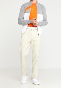 Veste zippée gris et blanc avec des accents orange portée sur un polo orange, associée à un pantalon crème et des baskets blanches. Textures lisses, coupe décontractée.