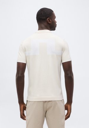 Mand med kort hår iført en beige kortærmet polo med et hvidt geometrisk mønster på den øvre ryg og matchende beige bukser.