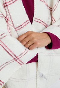 Coperta a quadri bianchi con linee rosse, in tessuto morbido e strutturato. Una mano con due anelli è visibile, che stringe il bordo della coperta.