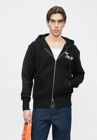 Čierna mikina na zips vyrobená z mäkkej tkaniny. Má predné vrecká, kapucňu so šnúrkou a biely logo detail na hrudi.