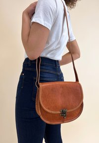 Brun läder crossbody-väska med en böjd klaff, metallspänne och synlig sömnad. Buren över en vit skjorta och jeans.