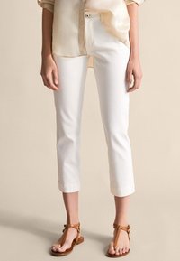 Białe, krótkie spodnie wykonane z bawełny, o prostych nogawkach, z bocznymi kieszeniami i podwiniętymi nogawkami. Połączone z brązowymi sandałami.