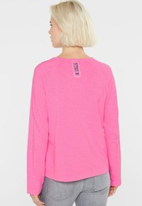 Frau mit kurzen blonden Haaren, die ein leuchtend pinkes Langarmhemd und helle graue Jeans trägt, von hinten zu sehen, mit sichtbarem Kleidungsetikett auf dem Rücken.