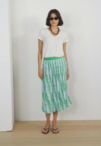 Bijela majica s v-neck izrezom u kombinaciji s plisiranom suknjom u zelenoj i plavoj tie-dye boji, koja ima elastičnu struk. Nosi se s crnim sandalama i sunčanim naočalama.