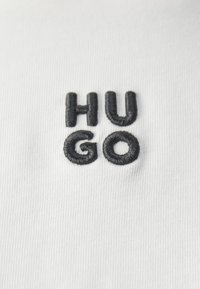 Hvit tekstil med svart brodert tekst som stavler "HUGO" i en moderne sans-serif skrifttype, med varierende bokstavhøyder og tykkelser.