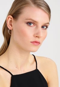 Zilveren creolen gedragen met een zwarte camisole, met een gladde textuur en cirkelontwerp; het model heeft steil haar en natuurlijke make-up.