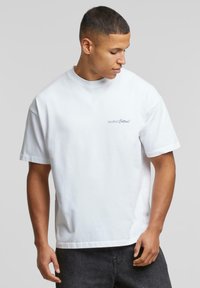 Hvid bomulds t-shirt med korte ærmer og rund hals, prydet med et lille blåt logo med teksten "Another Cotton!" på venstre side.