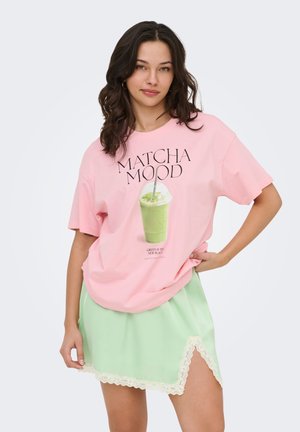 Vrouw draagt een oversized roze T-shirt met de tekst "MATCHA MOOD" en een afbeelding van een drankje, gecombineerd met een lichtgroene rok met kanten rand en een zijsplit.