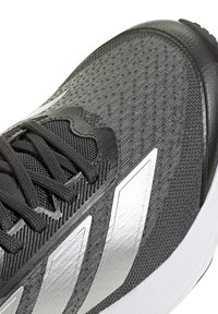 Gros plan d'une chaussure de sport noire et argentée avec une tige en mesh texturé et des lacets noirs sur fond blanc.