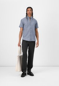 Kortermet blå chambray-skjorte, mørke bukser, svarte sko, holder en beige totebag. Nøytral bakgrunn, stående positur.