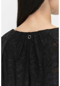 Svart blus med texturerad blommig brodyr och plisserad design på baksidan, med en metallring som stänger vid halslinningen.
