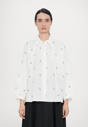Cămașă albă cu broderie florală verde, nasturi pe față, guler întins, mâneci lungi și voluminoase, și o croială lejeră.