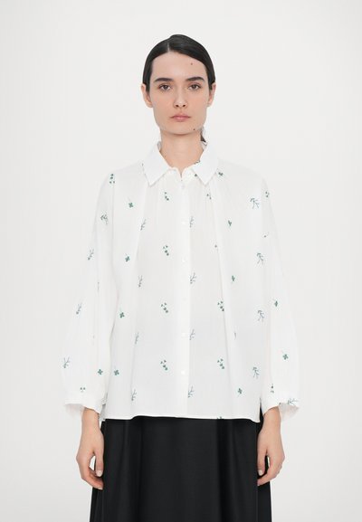 Chemise blanche avec broderie florale verte, patte de boutonnage à l'avant, col évasé, manches longues et volumineuses, et coupe décontractée.