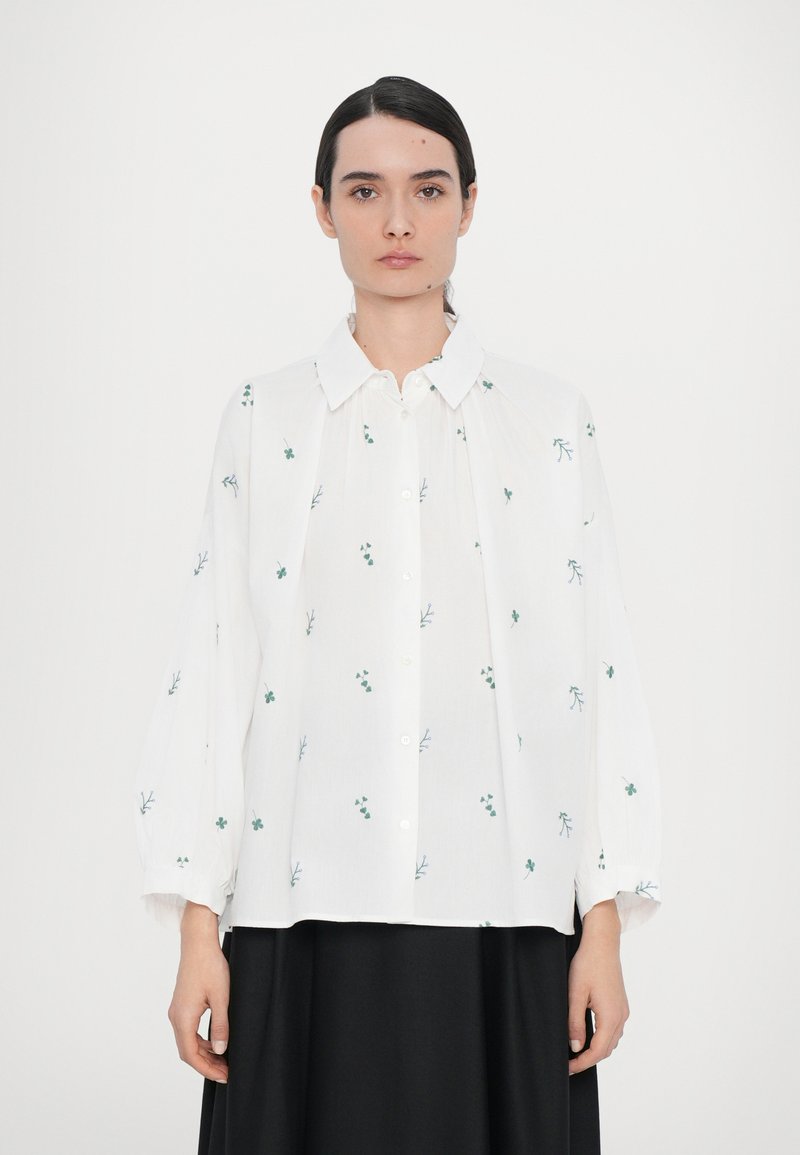 Chemise blanche avec broderie florale verte, patte de boutonnage à l'avant, col évasé, manches longues et volumineuses, et coupe décontractée.
