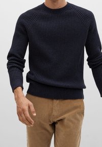 Homme portant un pull en maille côtelée bleu marine foncé et un pantalon en velours côtelé marron clair, debout avec un bras légèrement plié.