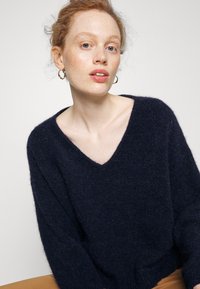 Pull en maille duveteuse bleu marine avec un col en V et une coupe ample. Doté de manches longues et larges ainsi que d'un ourlet côtelé. Texture douce avec un léger éclat.