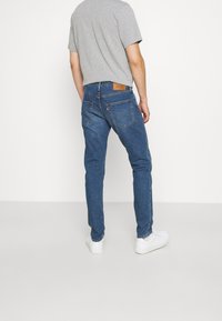 Blå denimjeans med smal passform, med en brun läderlapp på bakre midjan och en enkel grå t-shirt. Vita sneakers synliga.
