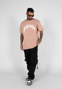 Lys rosa t-shirt med hvid grafisk tekst, parret med løse sorte cargo-bukser med lommer og stropper. Sko med sort og hvidt accent.