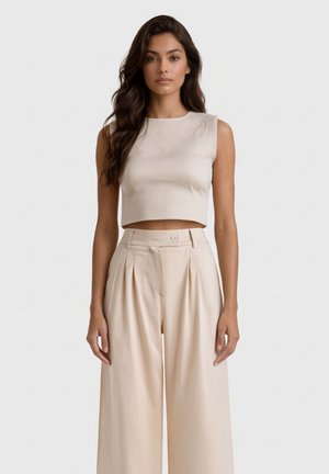 Donna con lunghi capelli scuri indossa un crop top beige senza maniche e pantaloni beige a gamba larga e vita alta, in piedi davanti a uno sfondo chiaro uniforme.