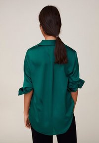 Woman with dark hair tied in a low ponytail wearing a green satin long-sleeve shirt with rolled-up sleeves, seen from the back.