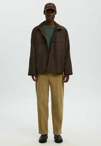 Hombre con chaqueta marrón, suéter verde, pantalones de pana beige, botas negras y gorra marrón con texto blanco, de pie contra un fondo claro.