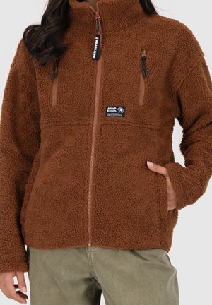 Brun fleecejakke med lynlås foran, to lomme med lynlås på brystet, et mærkepatch og sidelommer, båret med grønne bukser, hånd i lommen.