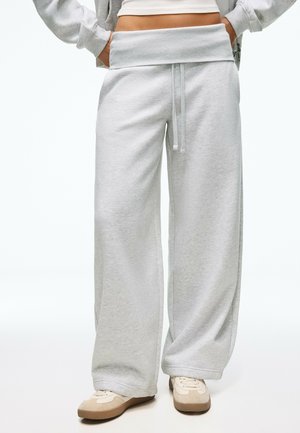 Personne portant un pantalon de survêtement gris clair à jambes larges avec taille à cordon, un crop top blanc, une veste grise et des baskets beiges. Mains dans les poches.