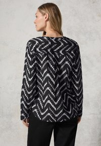 Blusa a righe zigzag bianche e nere, maniche lunghe, orlo arrotondato, tessuto liscio con una leggera lucentezza, vista posteriore che rivela il dettaglio della cucitura del collo.