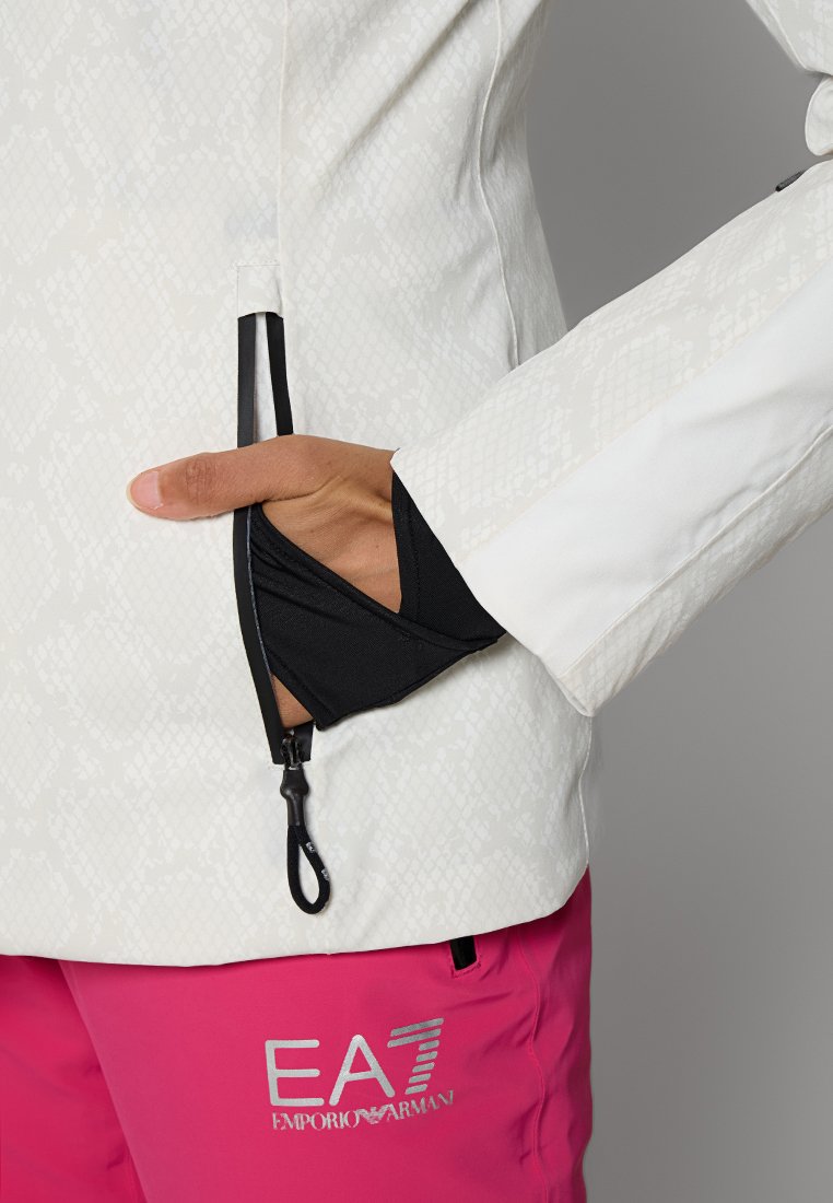 Weißes Jackett mit Schlangenhautmuster, ausgestattet mit einer schwarzen Reißverschlusstasche und einem schwarzen Innenfutter. Rosa Hose mit silbernem Logo-Detail.